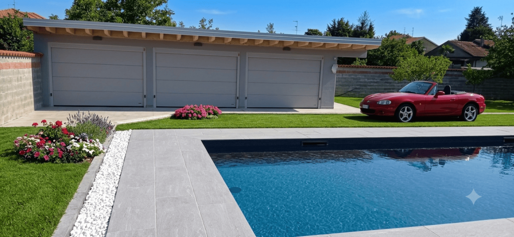 Nell'immagine si vede un bel giardino decorato con erba e aiuole colorate con fiori ed una piscina. Il cielo limpido riflette l'acqua della piscina con un bel colore azzurro. Sullo sfondo si vede una nuova auto cabriolet rossa ed un edificio adibito a garage con ben tre portoni sezionali bianchi.
