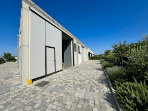 Nell'immagine si vede un moderno capannone artigianale edificato in una zona rurale con ampia vegetazione verdeggiante. L'ingresso al magazzino è possibile grazie ad un moderno portone a libro scorrevole, di colore grigio chiaro, con una porta pedonale inserita nel manto, utile come uscita di sicurezza.