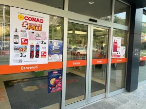 L'ingresso di questo supermercato Conad presenta una bussola con una coppia di porte automatiche sovrapposte, realizzate con. 2 ante fisse e due ante mobili gestite con automazioni ridondanti. 