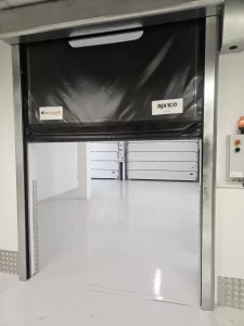 Nell'immagine si vede una porta rapida autoriparante con il telo di color nero, munito di finestrature trasparenti, per la compartimentazione dei reparti.In fondo si vede una coppia di portoni sezionali per la chiusura esterna.