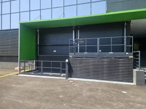 L'immagine mostra l'accesso delle merci del magazzino. Le merci vengono scaricate a terra su una piattaforma elevatrice che funge da montacarichi. Quando tutte le merci sono collocate sulla piattaforma, questa si alza al livello del magazzino. Si apre il portone sezionale e così si può trasportarla nei box affittati nello self storage.