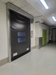 Nell'immagine si vede in primo piano una porta rapida autoriparante con il telo di color nero, munito di finestrature trasparenti, per la compartimentazione dei reparti. Il color nero è coordinato con i colori aziendali. 