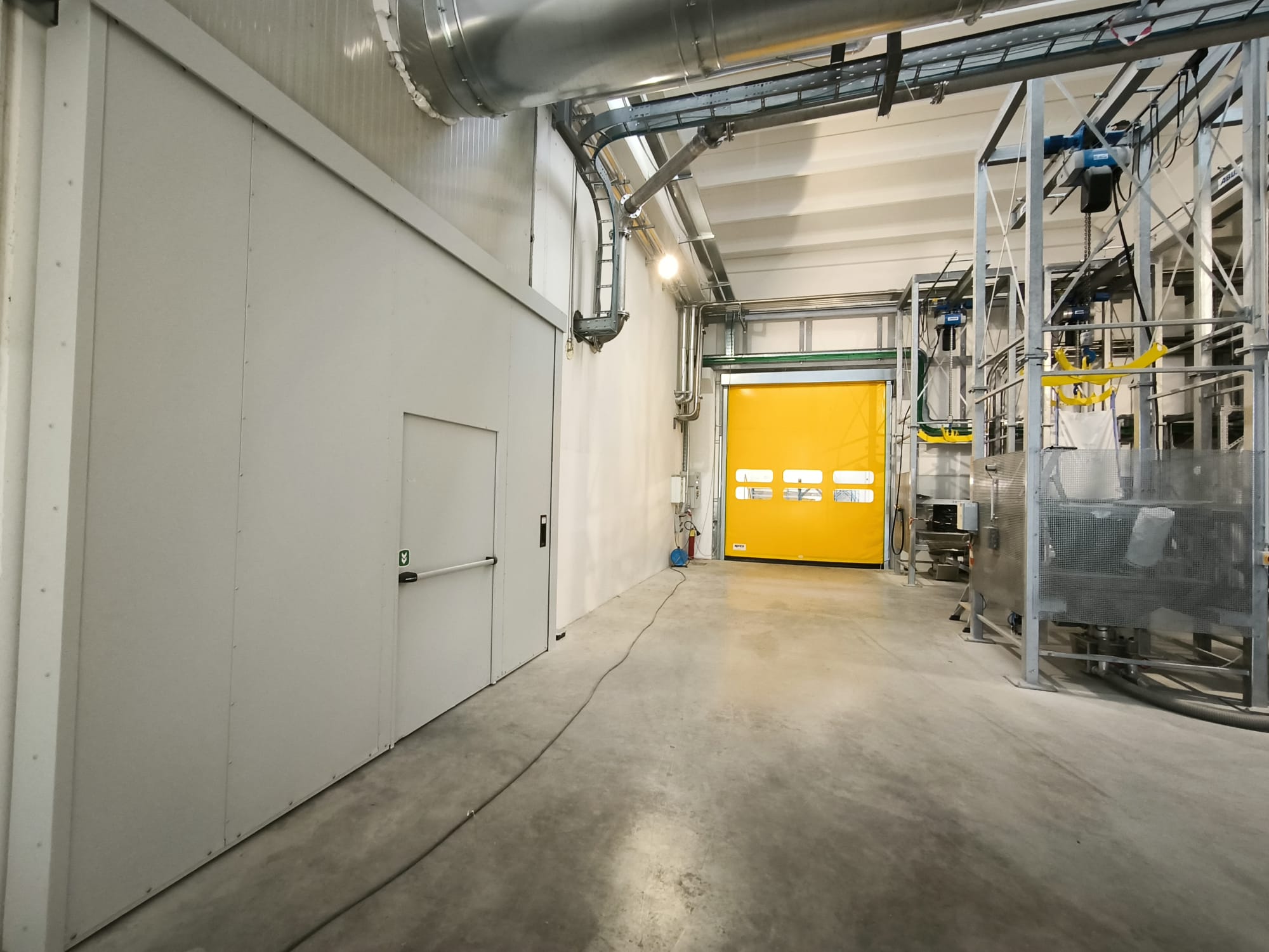 Nell'immagine si vede sulla sinistra un grande e largo portone scorrevole tagliafuoco bianco con inserito una porta a battente con maniglione antipanico usata come uscita di sicurezza. In fondo all'immagine si vede una porta con telo in pvc giallo ad avvolgimento rapido, con inserite 6 finestre in pvc trasparente