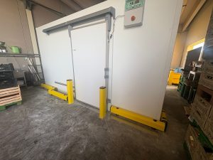 Nell'immagine si vede una porta automatica scorrevole per accesso ad ambiente con temperatura controllata, situato all'interno di un magazzino sanitario. Intorno al magazzino si vedono, a terra, i paracolpi ed i batticarrelli di colore giallo.