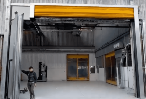 Questa immagine è una sequenza di quella precedente. La porta rapida è sempre impacchettata in alto. Si vede un tecnico che, sulla sinistra, ha aperto e fatto ruotare a 90° il montante verticale della porta insieme con l'anta del portone a libro. In questo modo ora il passaggio è più largo.