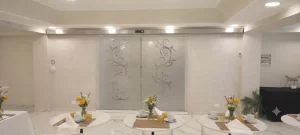 Nell'immagine si vede una porta automatica a due ante mobili tutto vetro con eleganti cristalli satinati bianchi finemente decorati. La porta è installata all'ingresso della sala da pranzo di un importante centro congressuale.