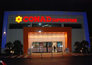 Nell'immagine ripresa in orario serale si vede l'ingresso di un grande supermercato Conad. L'ingresso è dotato di una coppia di porte automatiche con due ante fisse e due mobili, tutte dotate di sistemi a sfondamento integrale. Intorno e sopra le ante si vedono delle vetrate e dei sopraluce in eleganti cristalli satinati bianche.