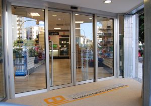 Nell'immagine si vede una porta automatica a due ante fisse e due mobili con sistema a sfondamento integrale e con serramenti color silver, installata all'ingresso di un grande negozio specializzato in prodotti e sistemi informatici.