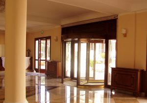 Nell'immagine si vede una elegante e prestigiosa porta automatica rotante con struttura in legno, installata nella hall di un grande albergo in edificio d'epoca.