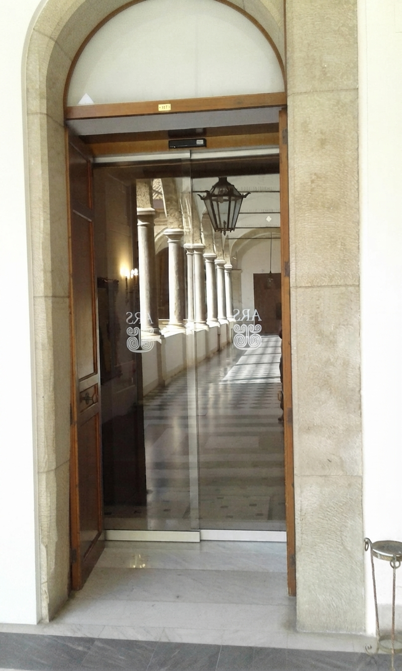 Porta automatica con due ante mobili in cristallo installate nel Palazzo dei Normanni. Attraverso i cristalli si vede l'elegante colonnato interno.