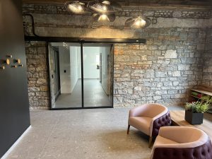 L'immagine mostra una elegante porta automatica tutto vetro all'interno della hall di un albergo. Nella hall si vedono eleganti poltrone, eleganti lampade e muri decorate con pietre carsiche.