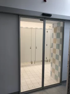 In questa foto si vede una pratica porta automatica con due ante in cristallo temperato ed un sottile serramento PAM 16 attraverso cui si accede al locale con le docce di una piscina o di una palestra.