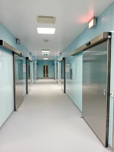 L'immagine mostra il corridoio di un reparto ospedaliero con un pavimento chiaro e muri colorati in azzurro. Gli ingressi alle varie stanze e laboratori sono dotate di porte automatiche a tenuta semplice con ante in acciaio.