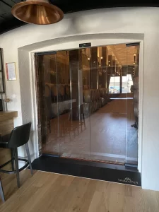 Una elegante porta automatica con due ante mobili e due ante fisse in cristallo temperato. E' l'interno di un elegante bar con in terra un parquet di vero legno ed eleganti lampade.