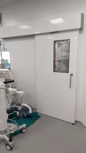 L'immagine mostra una porta automatica scorrevole a tenuta ermetica che da accesso alla sala operatoria. La porta è corredata da maniglione di sblocco di emergenza e visiva trasparente complanare.