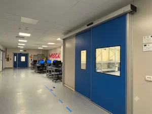 L'immagine in primo piano mostra una porta automatica con due ante telescopiche blu e visive trasparenti che danno accesso al magazzino interno di un'azienda di componenti sanitari.