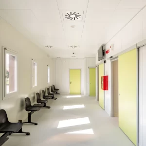 Nell'immagine si vede il corridoio di una sala d'attesa in una struttura sanitaria, con una serie di porte automatiche scorrevoli e porte a battente, che danno accesso ai reparti di visite mediche specialistiche.