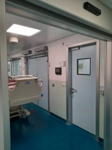 Nell'immagine si vede il corridoio con una barella ed una serie di porte automatiche a battente che danno accesso ai vari reparti di visite specialistiche. Le ante delle porte automatiche sono tutte di colore azzurro. Una delle porte e dotata di visiva complanare. 