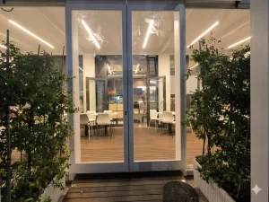 Nell'immagine si vede la veranda di un moderno ristorante. L'ingresso avviene grazie ad una porta automatica con due ante fisse e due ante mobili con sistema di sfondamento per vie di fuga, montate su serramenti a norma di color bianco.