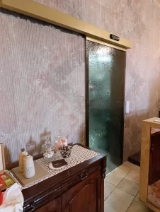 Nell'immagine si vede in primo piano una elegantissima porta scorrevole con un'anta mobile scorrevole filo muro. L'anta è tutto vetro con un cristallo satinato color verde giada. Posizionata all'interno di un bar, accompagna con eleganza gli ospiti nelle toelette.