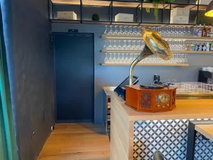 Nell'immagine si vede in primo piano il bancone di un bar decorato con un vecchio grammofono in legno ed ottone. In secondo piano si vede una porta scorrevole ad un'anta con cristallo fumè che porta ad un magazzino di servizio.