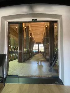 Nell'immagine si vede una porta scorrevole tutto vetro con due ante scorrevoli e due ante fisse che portano all'interno di una cantina vitivinicola. Attraverso la porta si vedono lunghi corridoi con antiche botti dove riposano vini e grappe selezionate.
