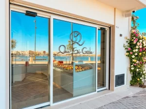 In questa immagine si vede l'ingresso automatico di una gelateria e bar situata in un paese di mare. Si notano due porte automatiche scorrevoli divise da un'anta fissa. Eleganti serramenti di color bianco incorniciano i banchi interni piedi di dolci deliziosi.