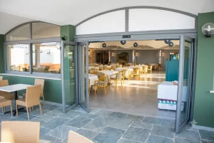 Nell'immagine si vede la veranda e l'interno di un ristorante con tavoli imbanditi. L'ingresso al ristorante è gestito da una porta automatica scorrevole con due ante fisse e due ante mobili e sistema di sfondamento integrale. Nell'immagine si vedono le ante nella posizione di sfondamento totale.