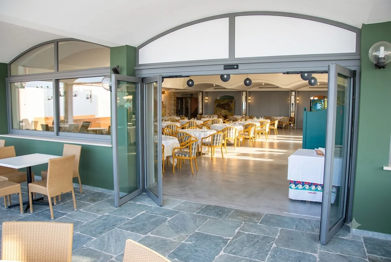 Nell'immagine si vede la veranda e l'interno di un ristorante con tavoli imbanditi. L'ingresso al ristorante è gestito da una porta automatica scorrevole con due ante fisse e due ante mobili e sistema di sfondamento integrale. Nell'immagine si vedono le ante nella posizione di sfondamento totale.