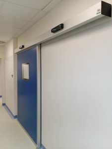 Porta scorrevole a chiusura ermetica installata all'ingresso di sala operatoria. Pannello blu con inserita visiva trasparente complanare.