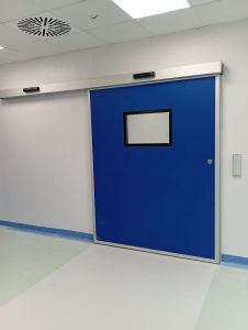 Porta scorrevole a chiusura ermetica installata all'ingresso di sala operatoria. Pannello blu con inserita visiva trasparente complanare. Cassonetto con automazione e radar attivazione aperture e chiusure.