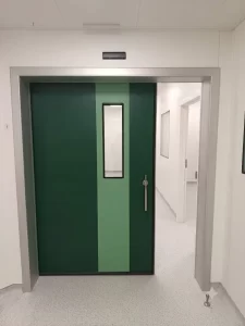 Nell'immagine si vede una porta automatica scorrevole a tenuta ermetica, di colore verde, parzialmente aperta. L'immagine è presa dall'interno della sala operatoria, pertanto si intravede il corridoio esterno con le aperture dei locali attigui. Sull'anta si vede il maniglione di sicurezza per l'apertura manuale e la visiva verticale per vedere all'interno. Sulla parete il pulsante a sfioro per l'apertura automatica.