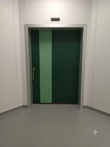 Nell'immagine si vede una porta automatica scorrevole a tenuta ermetica, di colore verde, chiusa. Sull'anta si vede il maniglione di sicurezza per l'apertura manuale. Sopra la porta, sulla parete, si vede il radar che comanda l'apertura automatica.