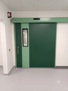 Nell'immagine si vede una porta automatica scorrevole a tenuta ermetica, di colore verde. Sull'anta si vede il maniglione di sicurezza per l'apertura manuale e la visiva verticale per vedere all'interno. Sulla parete il pulsante a sfioro per l'apertura automatica. Il cassonetto è colorato di verde.