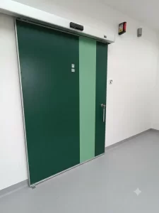 Nell'immagine si vede una porta automatica scorrevole a tenuta ermetica, di colore verde. Sull'anta si vede il maniglione di sicurezza per l'apertura manuale. Sulla parete il pulsante a sfioro per l'apertura automatica.