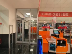 Nell'immagine si vede l'uscita di un supermercato con il primo piano le casse automatiche e, di lato, la prima parte di una bussola con porte automatiche. La prima porta automatica è una porta scorrevole rettilinea telescopica con due ante mobili e una fissa sulla quale vanno a sovrapporsi le due mobili. La scelta della porta telescopica è dovuta allo scarso spazio disponibile lateralmente nel corridoio di uscita.
