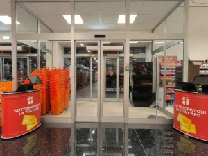 Nell'immagine si vede l'ingresso di un supermercato con una bussola composta da una coppia di porte automatiche scorrevoli contrapposte. Le porte sono realizzate ognuna da due ante mobili ed due anti fisse. La bussola con le due porte distanziate permette di creare una zona cuscinetto che mitica la dispersione termica tra interno ed esterno, favorendo il risparmio energetico.