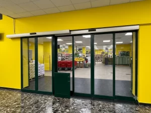 Nell'immagine si vede l'ingresso di un supermercato. I colori giallo dei muri e verde dei serramenti rispecchiano i colori aziendali del logo. L'ingresso è composto da una coppia di porte automatiche scorrevoli con due ante mobili ciascuna più un'anta fissa centrale ed un'altra anta fissa laterale. Le porte automatiche sono a sfondamento integrale per vie di fuga.