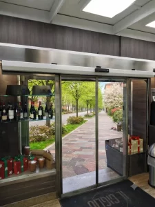 Nell'immagine si vede l'ingresso di un negozio di alimentari città. L'ingresso è gestito da una porta automatica con due ampie ante mobili e due ante fisse. L'automazione è gestita da un sistema ridondante che garantisce il funzionamento come vie di fuga.