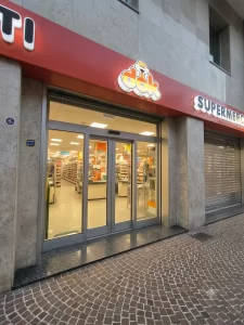Nell'immagine si vede l'ingresso di un supermercato della catena DOK con al centro una porta automatica scorrevole a due ante, due sezioni vetrate ai lati ed un sopraluce in vetro.