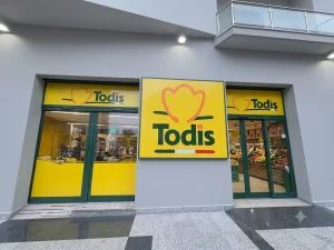 Nell'immagine si vedono l'ingresso e l'uscita da un supermercato di città della catena Todis. I serramenti sono di colore verde coordinati come il logo aziendale. L'entrata è gestita da una porta telescopica a tre ante mente l'uscita è gestita da una larga anta mobile più una fissa. La parete esterna tra le due porte automatiche ed i sopraluce sono decorati con il logo aziendale.