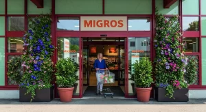 Nell'immagine si vede una cliente di un supermercato uscire con facilità spingendo un carrello della spesa colmo di merci, attraverso una porta automatica scorrevole. La porta è una porta scorrevole automatica a due ante fisse e due mobili con sistema di sfondamento integrale, utile come via di fuga in caso di emergenza.
