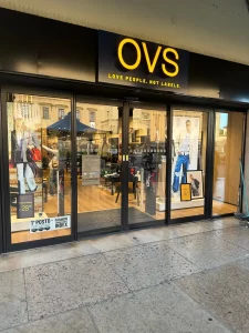 L'immagine mostra la vetrina ed una porta scorrevole a due ante mobili scorrevoli dietro le vetrine, di un negozio della OVS aperto nella galleria di un centro commerciale. La scritta OVS sopra la porta automatica e le vetrine è nel consueto colore giallo aziendale. La foto è stata realizzata con il negozio operativo e con già gli scaffali pieni di merci.