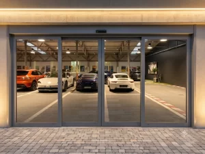 Nell'immagine si vede l'ingresso di una show room Porsche. All'interno diverse autovetture parcheggiate in esposizione. Per l'ingresso si vede una porta automatica con due ante scorrevoli e due ante fisse.