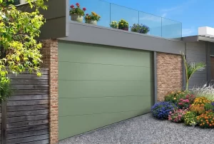 L'immagine mostra un garage con un vialetto d'ingresso inclinato. Il manto della porta del garage, di un gradevole colore verde salvia, presenta l'ultimo pannello inferiore sagomato a trapezio, in modo da essere complanare con l'inclinazione del vialetto. La guarnizione inferiore presente sull'ultimo pannello permette di isolare il pavimento del garage dal vialetto d'ingresso.