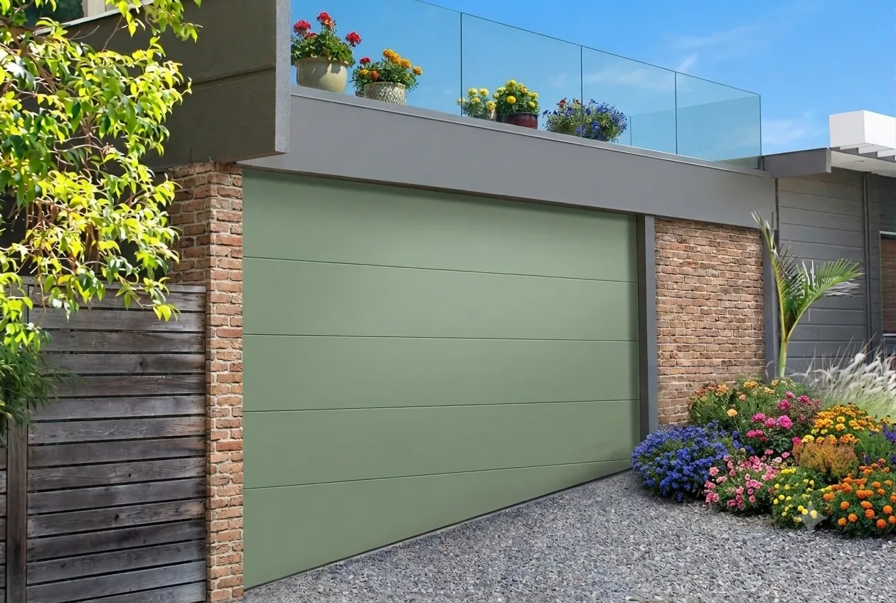 L'immagine mostra un garage con un vialetto d'ingresso inclinato. Il manto della porta del garage, di un gradevole colore verde salvia, presenta l'ultimo pannello inferiore sagomato a trapezio, in modo da essere complanare con l'inclinazione del vialetto. La guarnizione inferiore presente sull'ultimo pannello permette di isolare il pavimento del garage dal vialetto d'ingresso.