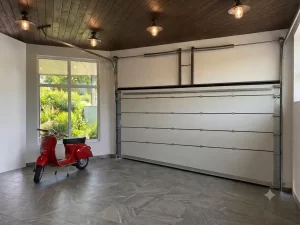 L'immagine mostra l'interno del garage caratterizzato da una forte inclinazione del pavimento all'ingresso. Per questo motivo l'ultimo pannello inferiore non ha la classica forma rettangolare, bensì a trapezio, in modo da adattarsi perfettamente in modo complanare al pavimento. Nel garage, caratterizzato da una elegante pavimentazione grigia e da un caldo soffitto ricoperto da pannelli di legno, si vede una iconica motocicletta Vespa di color rosso.