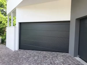 Nella foto si vedono gli ingressi dei garage di una casa moderna gestiti da due portoni sezionali color antracite. In questa foto si vede il portone sezionale di sinistra con l'ultimo pannello inferiore a forma di trapezio in modo da adattarsi in modo complanare alla pavimentazione del vialetto particolarmente inclinata e chiudere in modo perfetto.