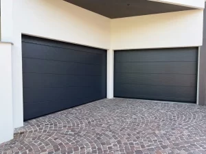 Nella foto si vedono gli ingressi dei garage di una casa moderna gestiti da due portoni sezionali color antracite. In questa foto i due portoni sezionali sono chiusi. Il garage di sinistra è caratterizzato da una pavimentazione all'ingresso particolarmente inclinata. Per questo motivo l'ultimo pannello inferiore non ha la tradizionale forma rettangolare, bensì a trapezio in modo da adattarsi in modo complanare alla pavimentazione del vialetto e chiudere in modo perfetto.