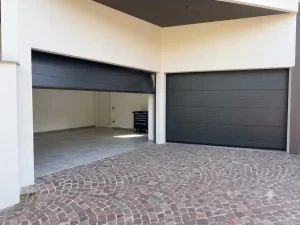 Nella foto si vedono gli ingressi dei garage di una casa moderna gestiti da due portoni sezionali color antracite. In questa foto il portone sezionale di sinistra è parzialmente aperto mentre quello di destra è chiuso. Si vede che l'ultimo pannello inferiore non ha la tradizionale forma rettangolare, bensì a trapezio in modo da adattarsi in modo complanare alla pavimentazione del vialetto inclinata e chiudere in modo perfetto. Il garage di sinistra è infatti caratterizzato da una pavimentazione all'ingresso particolarmente inclinata.